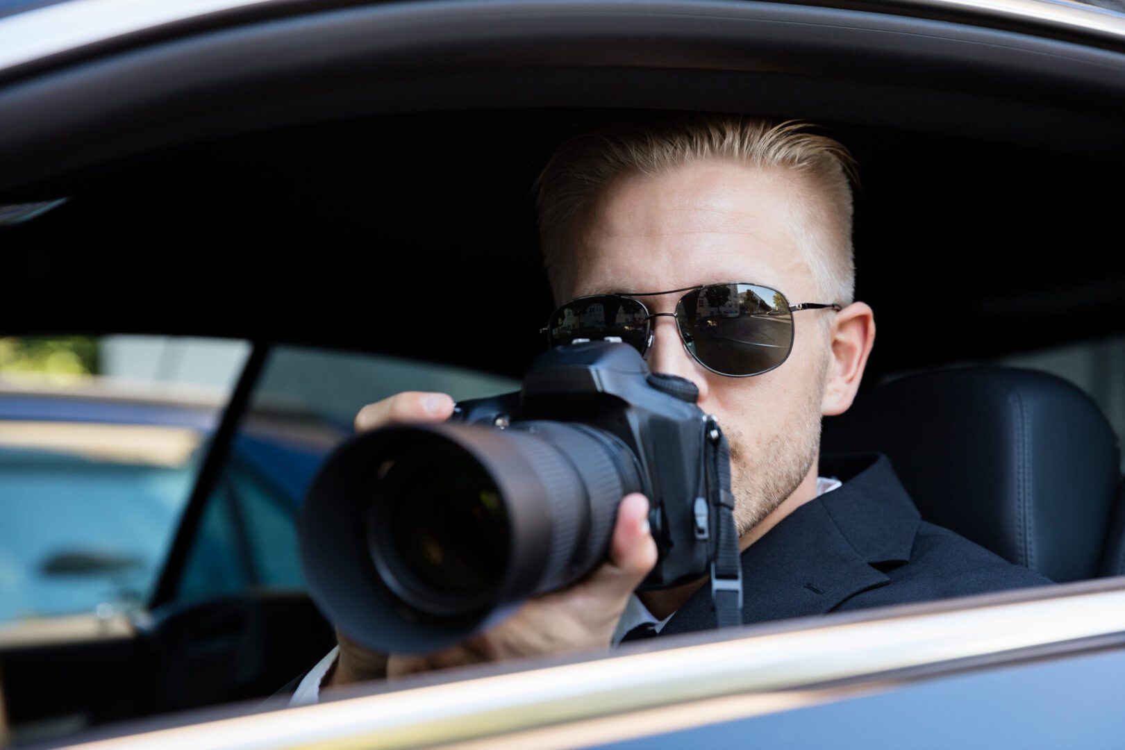A man in sunglasses holding a camera while sitting inside of a car.